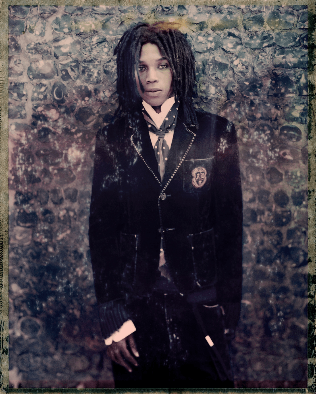 A portrait of a person with dreadlocks wearing a dark school uniform-style blazer with a crest patch, over a light shirt and dark tie, standing against a textured stone wall.