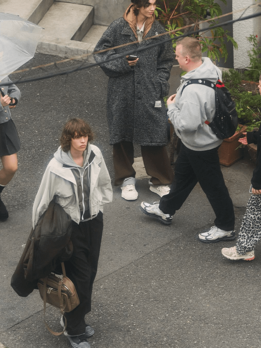 A street-style photograph showing several people outdoors on a paved walkway. In the foreground, a person wearing a light gray zip-up hoodie and black pants carries a large dark tote bag.