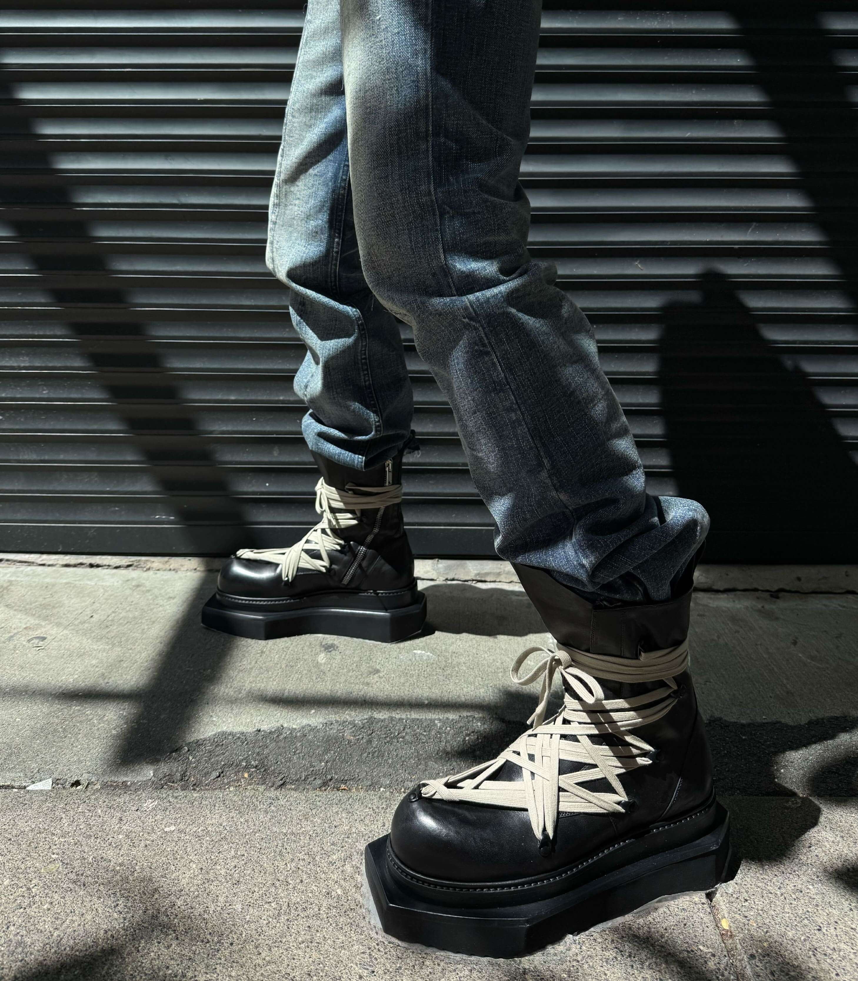 Person wearing Rick Owens black platform boots with beige laces against a metallic wall.