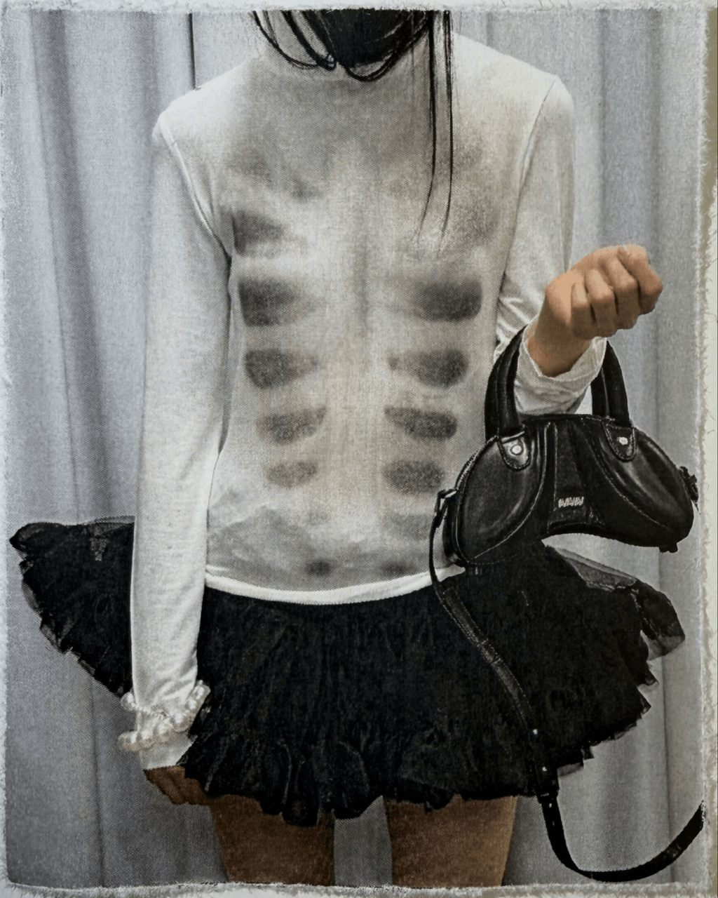 A person wearing a white long-sleeve top with a black ribbed ribbed pattern across the chest, paired with a black ruffled mini skirt and holding a black handbag, set against a gray curtain backdrop.