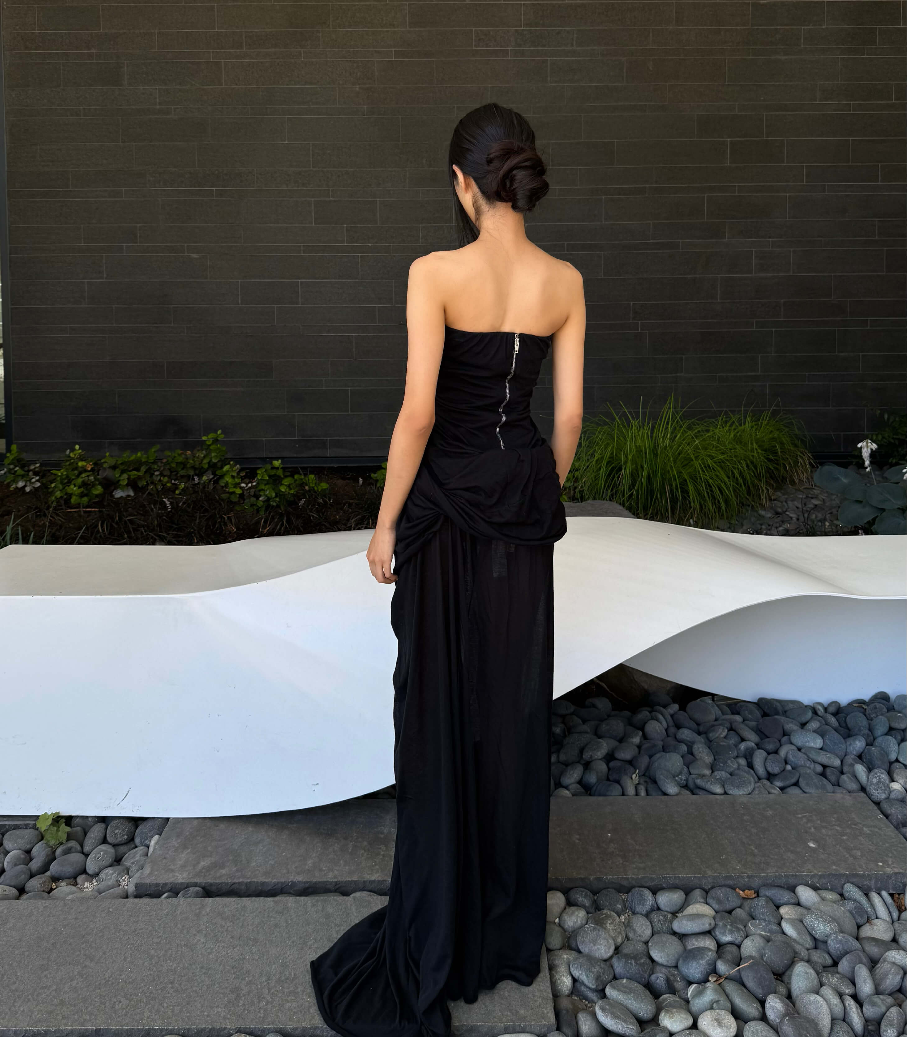 Woman in a Rick Owens black strapless gown standing in front of a modern sculpture with a dark wall and plants in the background.