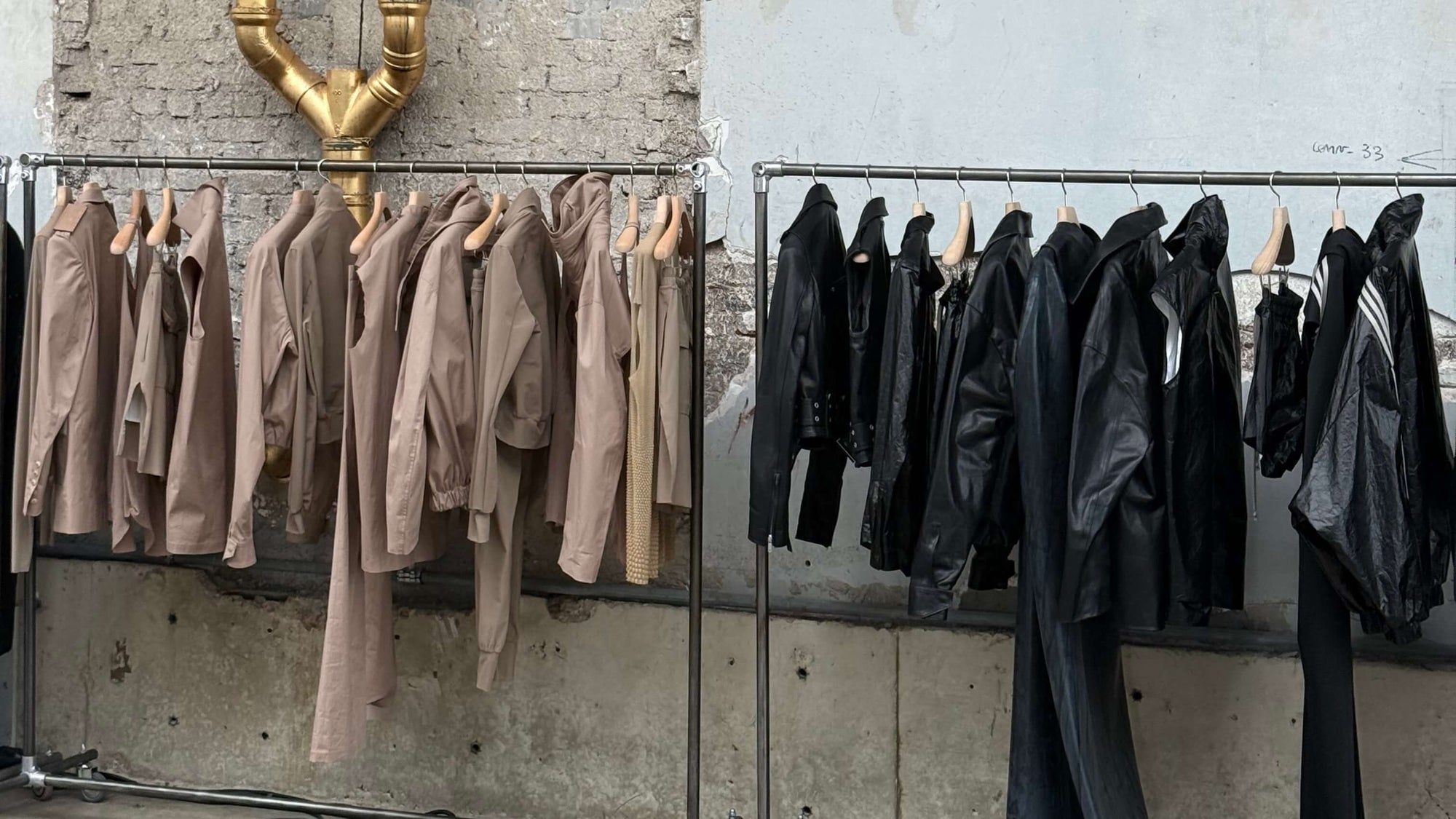 Two clothing racks displaying garments in a warehouse-style showroom with exposed brick and concrete walls. The left rack holds light beige/tan jackets, and the right rack holds black leather jackets and a long black coat.